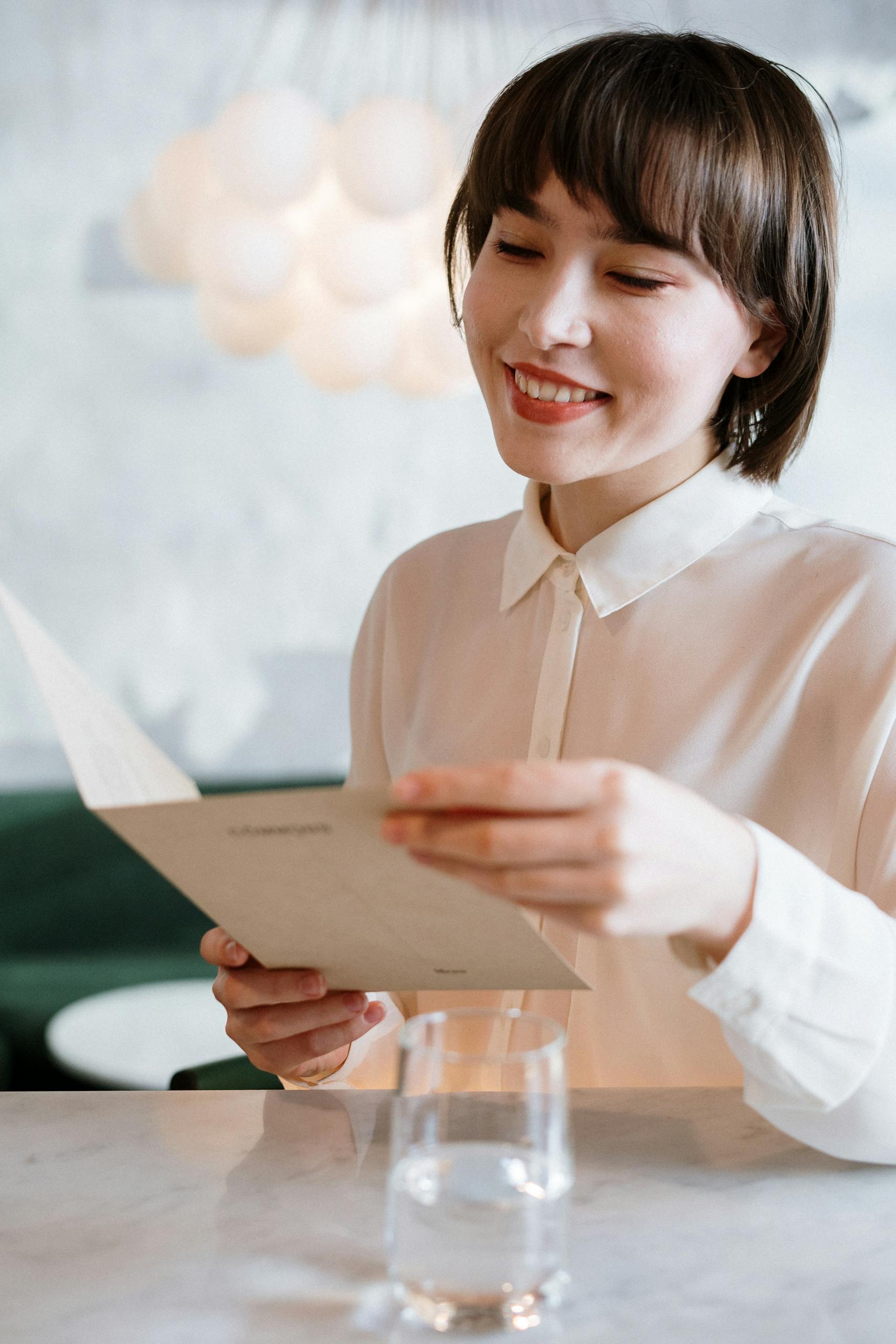 A smiling young woman in a white shirt reads a menu in a stylish cafe environment.